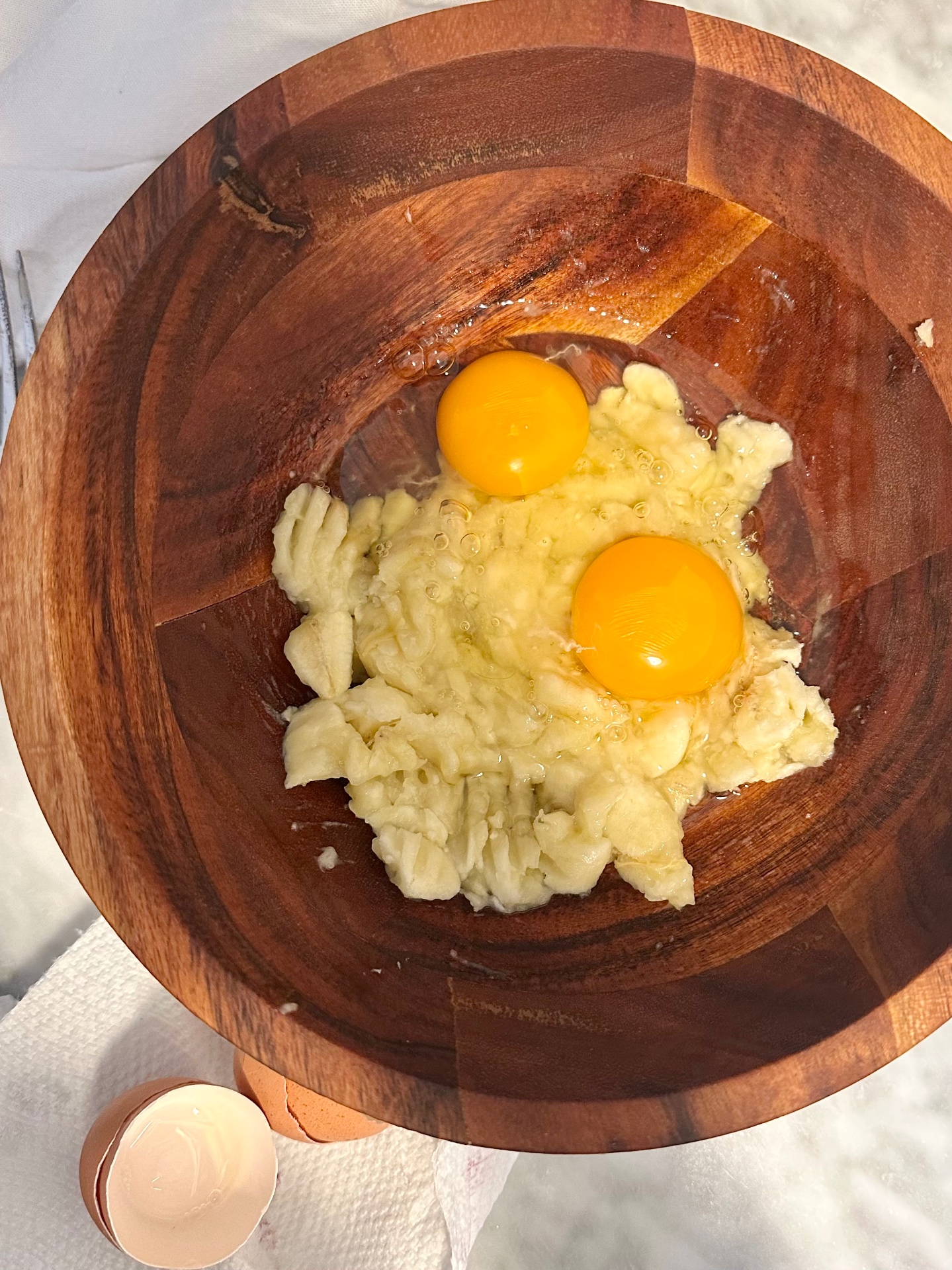 Mashed banana with two cracked eggs in a wooden mixing bowl