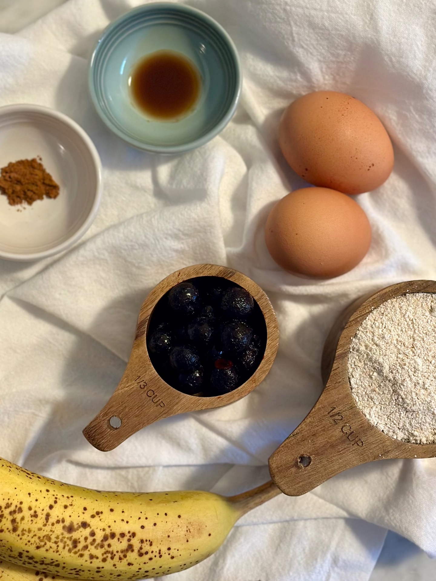Recipe ingredients arranged on white linen: ripe bananas, eggs, blueberries, oat flour, vanilla, and cinnamon