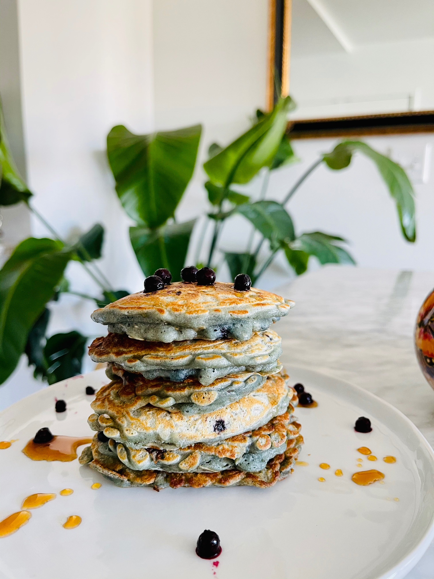 A tall stack of homemade banana blueberry pancakes finished with blueberries and a light maple syrup drizzle