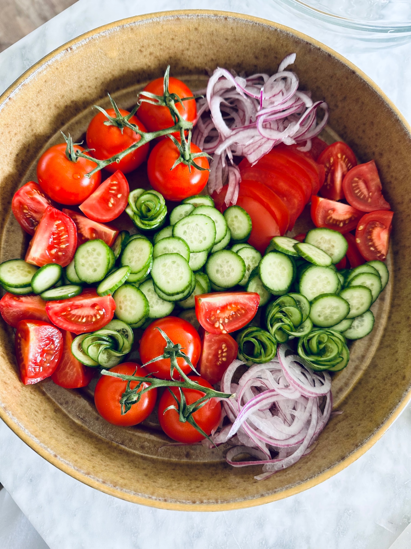 A shallow ceramic bowl arranged with vine tomatoes, cucumber slices, thinly shaved red onion, and fresh basil on marble