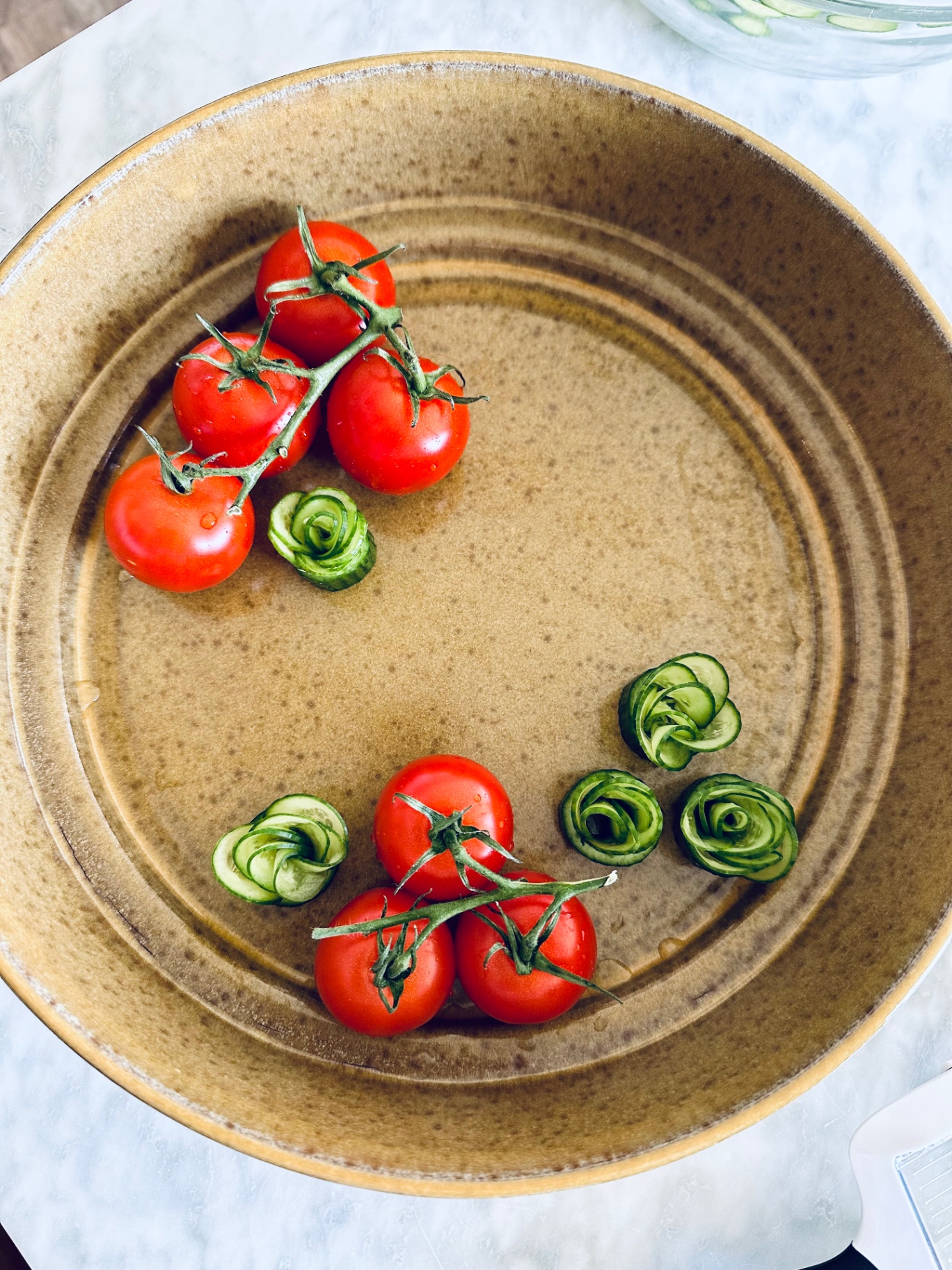 Tomatoes and rolled cucumber ribbons arranged in an empty ceramic bowl, mid assembly