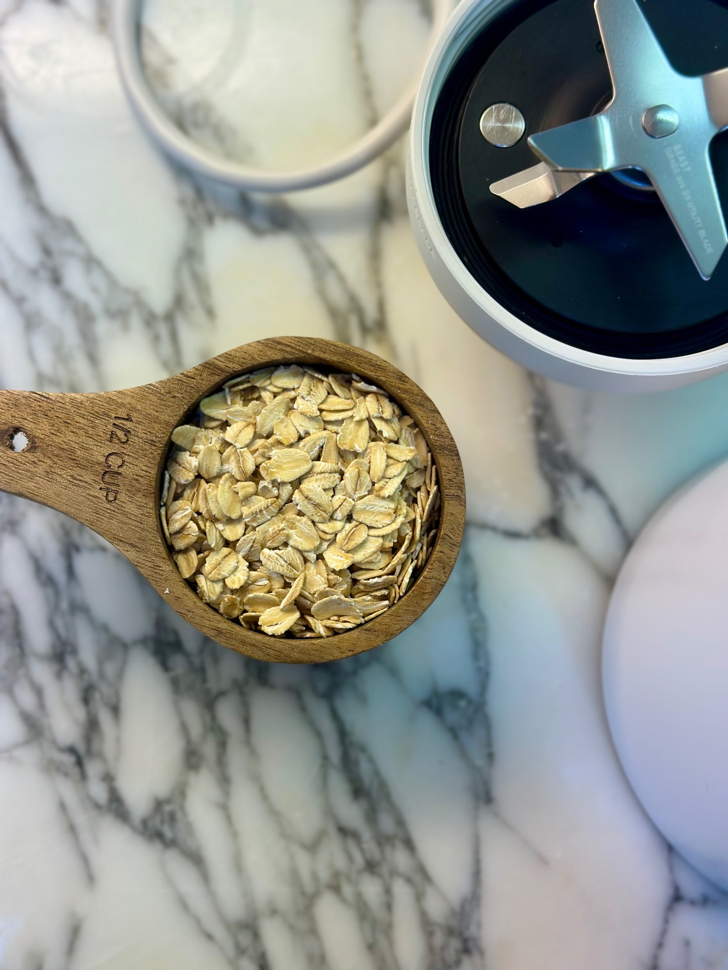 Oats in a wooden measuring cup beside an open blender on a marble counter